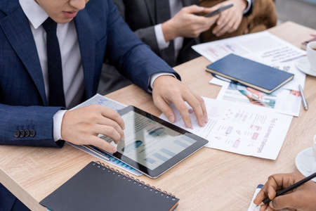 Contemporary economist in suit looking through charts and diagrams in tablet while sitting by table...