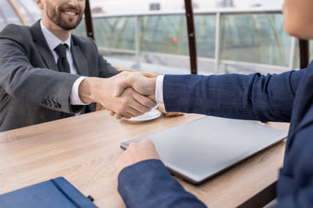Two young successful business partners in formalwear handshaking over table after negotiating and making dealの写真素材