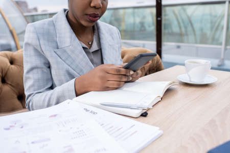 Hands of young African businesswoman scrolling in smartphone while sitting by table with papers and open notebookの写真素材