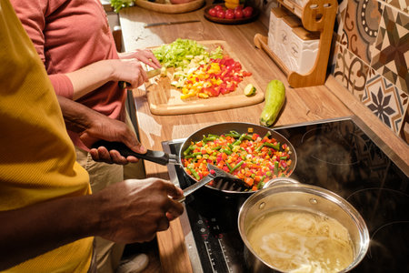 Young African man mixing chopped vegetables in frying-pan while standing by electric stove and cooking food with his wifeの写真素材
