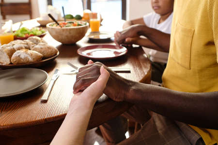Hands of African man, his Caucasian wife and their son during pray by table before having dinner or breakfastの写真素材