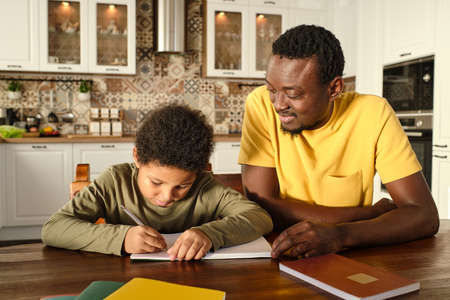 Happy young African man and his biracial son with pen and copybook carrying out homework by wooden tableの写真素材