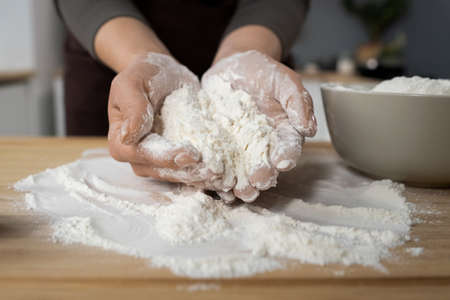 Hands of young female holding pile of sifted flour over wooden board while preparing homemade pastry in the kitchenの写真素材