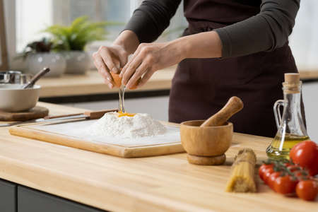 Hands of young woman breaking egg on top of pile of sifted flour on wooden board while making doughの写真素材