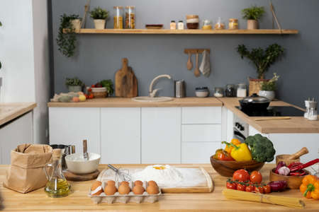 Interior of kitchen with wooden table, fresh vegetables, sifted flour, eggs, bottle of olive-oil and...
