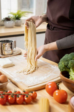 Hands of young woman holding boiled spaghetti over wooden board while preparing homemade food by kitchen tableの写真素材