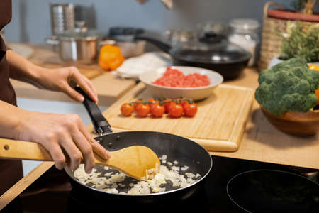 Hands of young woman mixing chopped onion in frying pan while standing by electric stove in the kitchenの写真素材