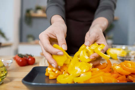 Hands of female putting chopped fresh yellow capsicum on tray while cooking vegetarian stew for dinner in the kitchenの写真素材