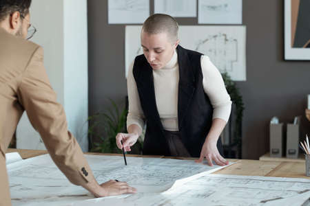 Confident businesswoman bending over table with blueprint while discussing sketch with young male...