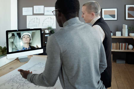 Two contemporary architects communicating to female builder on computer screen while standing by table with blueprintの写真素材