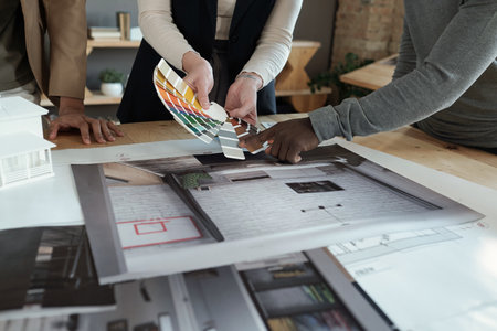 Hands of three intercultural coworkers standing by workplace with photos of apartment interiors...
