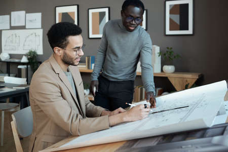 Two young intercultural businessmen discussing sketch of architecture on blueprint while one of them...