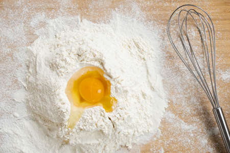 Flatlay of broken raw egg on top of pile of flour prepared for making homemade dough and whisk on...