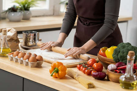 Hands of young female rolling homemade dough on wooden board while standing by kitchen table