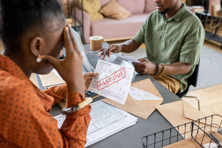 Young stressed female looking at unpaid bill and touching her head while sitting by table in front of man networkingの写真素材