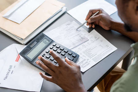 Hands of African man pressing buttons of calculator while checking data in financial papers and unpaid bills by tableの写真素材