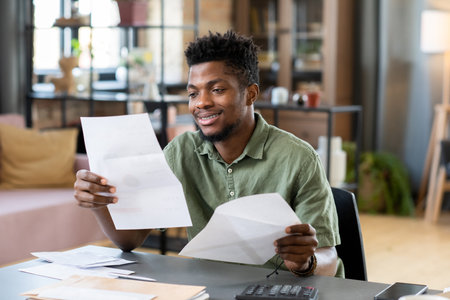 Young smiling African man looking through financial paper or paid tax payment form while sitting by tableの写真素材