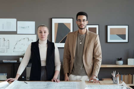 Two contemporary engineers standing by table with sketch and looking at you against grey wall with...