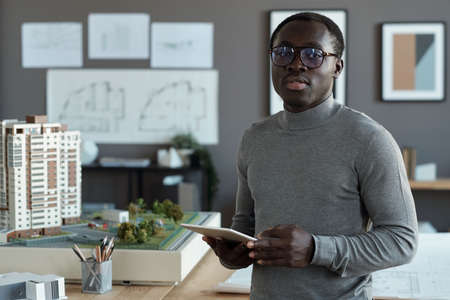 Professional engineer of African ethnicity scrolling in tablet while standing in office and looking for online sketchesの写真素材