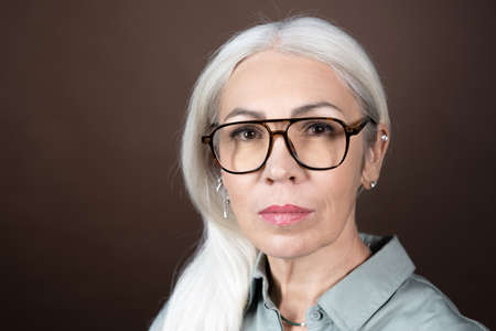 Close-up of mature woman in eyeglasses with long white hair looking at camera isolated on brown backgroundの写真素材