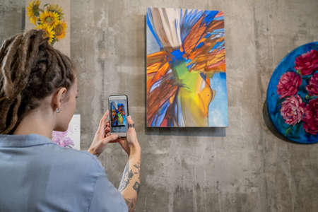 Contemporary young woman taking photo of abstract painting on wall while visiting exhibition in art gallery at leisureの写真素材