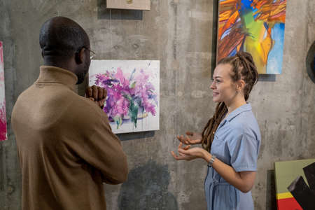 Young female guide describing one of paintings on walls to African man visiting modern art galleryの写真素材