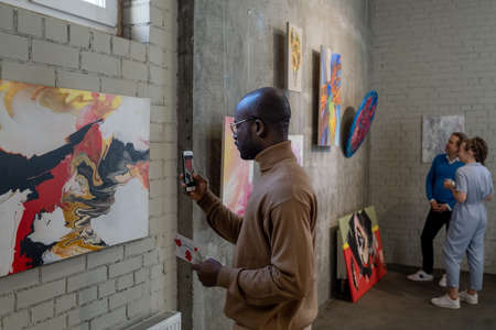 Young African male visitor of art gallery taking photo of painting on wall while enjoying exhibition of modern artistsの写真素材