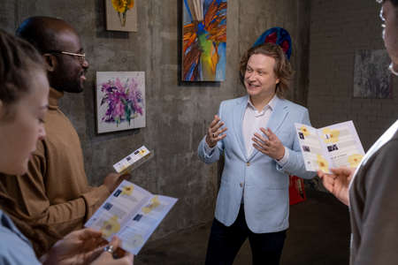 Happy elegant artist interacting with intercultural guests of exhibition of his artworks in galleryの写真素材