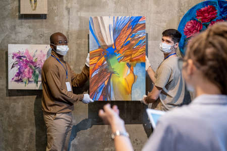 Two gallery workers in gloves and protective masks holding abstract painting and looking at female colleague while going to hang artwork on wallの写真素材