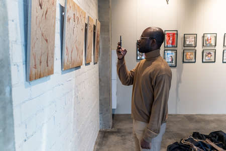 Young African man with smartphone taking photo of painting in museum while standing in front of wall with artwork collectionの写真素材