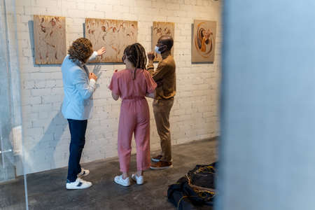 Young intercultural couple in masks listening to mature artist during presentation of his new artwork collection in galleryの写真素材