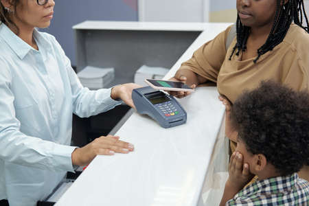 Young mother paying for dentistry service for her little son with toothache while holding smartphone over payment terminalの写真素材