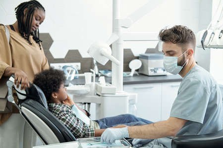 Male dentist taking hook and mirror for dental check-up out of metallic tray before examining oral cavity of little patientの写真素材