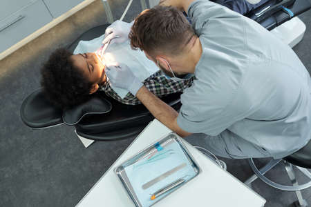 Young dentist in uniform and gloves using dental equipment during medical procedure while bending...