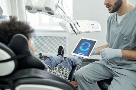 Gloved dentist in uniform showing human jaws and teeth on tablet screen to little patient while sitting in front of himの写真素材