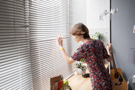 Sick young woman in dress looking through venetian blinds while bending by window of hospital wardの写真素材