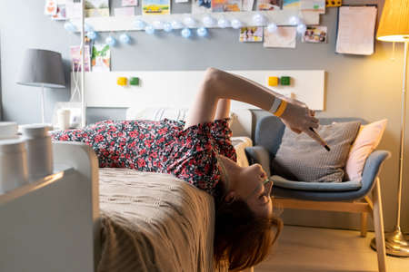Sick teenage girl in casual dress scrolling in smartphone while lying on bed in hospital ward of...