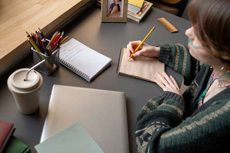 Creative teenager with pencil over page of notepad sitting by desk and thinking of new ideas while...