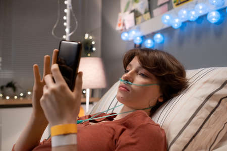 Brunette teenager with oxygen nose pipe texting in smartphone while lying on bed in hospital ward at...
