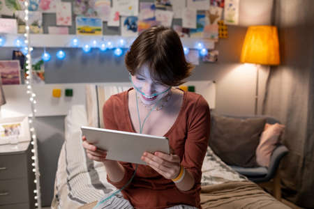 Cute happy teenager with tablet watching online video or communicating in online chat while sitting...