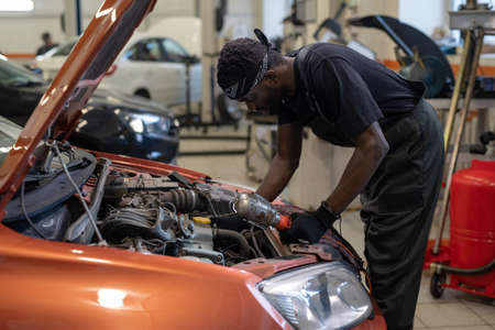 Young African man in workwear bending over open car hood while checking engine or repairing details...