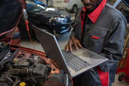 Part of repairman in workwear holding laptop while standing by car with open hood and consulting clients online in garageの写真素材
