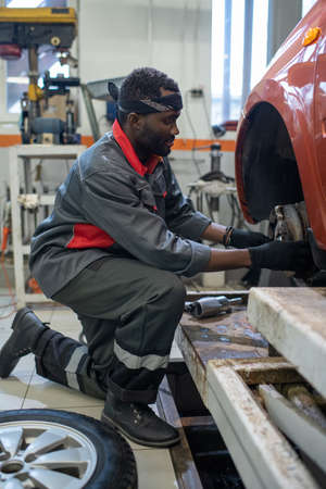 Young professional automechanic of African ethnicity in workwear and protective gloves changing...