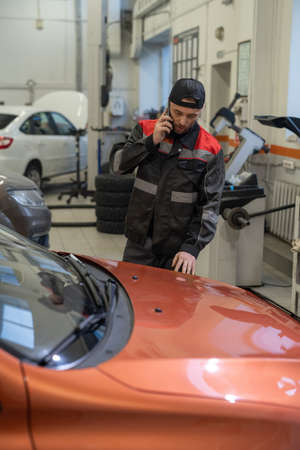 Young confident repairman speaking to client on mobile phone while standing by car after check-up...