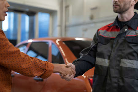 Handshake of young Hispanic female client and repairman in workwear standing against repaired car in garage or workshopの写真素材