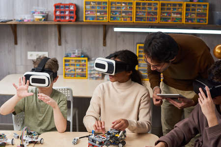 Group of children sitting at the table and projecting robots in vr glasses with teacher assisting them during robotics lessonの写真素材