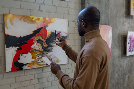 Rear view of African man using his mobile phone to make photo of artwork on the wall during...