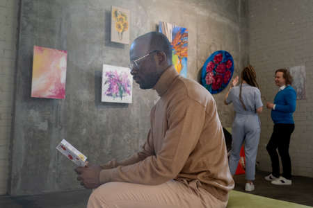 African serious man in eyeglasses reading a brochure while sitting in art gallery with other people...