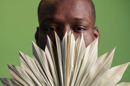 Part of face of young black man peeking out of white fan in front of camera while standing in...