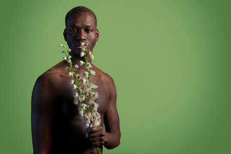 Young muscular African man with white flowers by his face and body standing in front of camera over...
